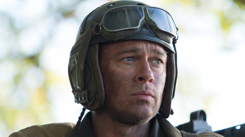 Don has his gloved hands clasped, a pilot's helmet with goggles resting on his head, looking into the distance pensively next to a machine gun in Fury