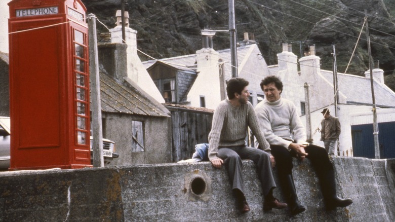 Peter Riegert as Mac sitting on a harbor wall beside a red telephone box in Local Hero