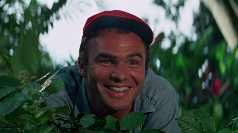 Burt Reynolds as Douglas Temple smiling while wearing a red baseball cap in Skullduggery