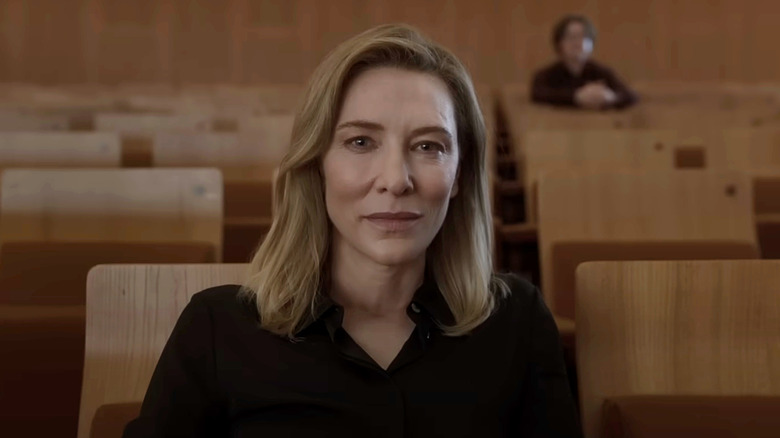 Cate Blanchett's Lydia Tár sits in a row of wooden seats looking straight into camera in Tár