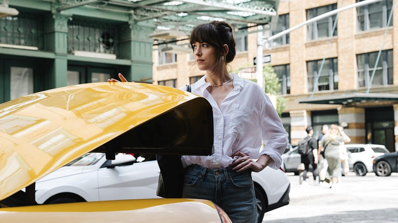 Dakota Johnson's Lucy wearing a white blouse and standing behind a cab with an open trunk in Materialists