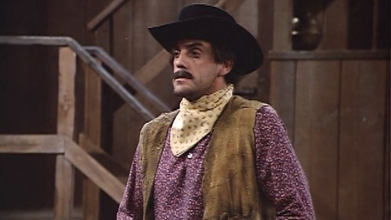 The Calico Kid (Christopher Lloyd) stands tall and serious in a cowboy hat, vest, and kerchief in front of a rustic wooden room in Best of the West
