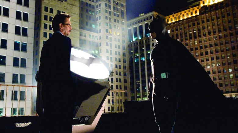 Gary Oldman's Jim Gordon standing with Christian Bale's Batman by the Bat-Signal in Batman Begins