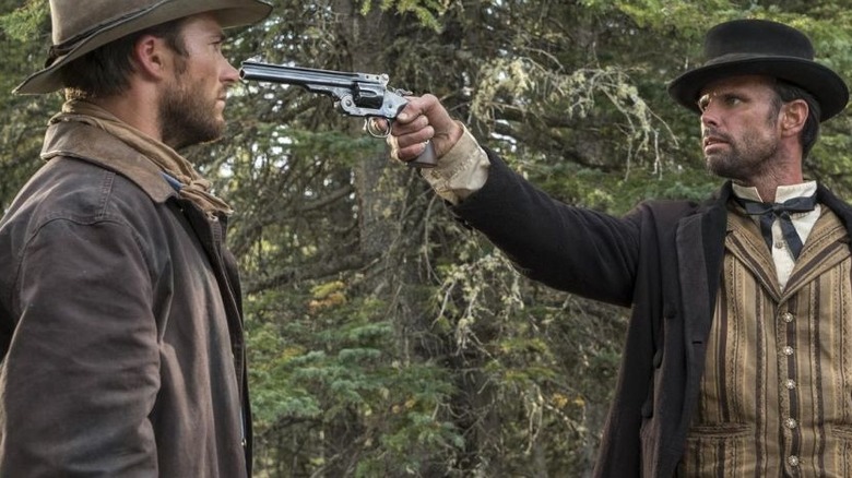 Ezra (Walton Goggins), mengenakan topi dan setelan bowler, mengarahkan revolver di koboi Jackson (Scott Eastwood) di tengah hutan selama adegan kebuntuan tegang ini dari Diablo
