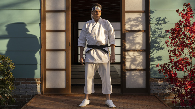 Daniel LaRusso standing in front of the Miyagi-Do dojo in a white gi and headband in Cobra Kai