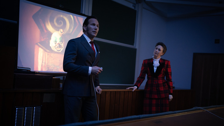 Ed and Lorraine Warren give a presentation in front of a projector screen showcasing a creepy clown doll in The Conjuring: Last Rites