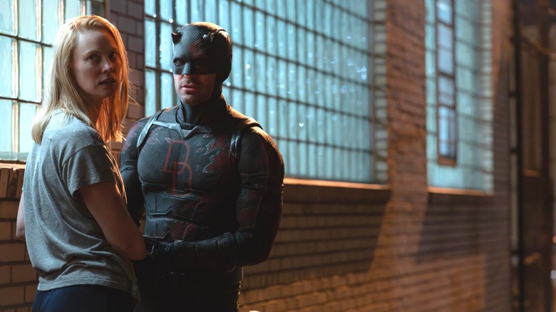 Charlie Cox as Matt Murdock in his superhero suit holding hands with Deborah Ann Woll as Karen Page by a wall of windows in Daredevil: Born Again