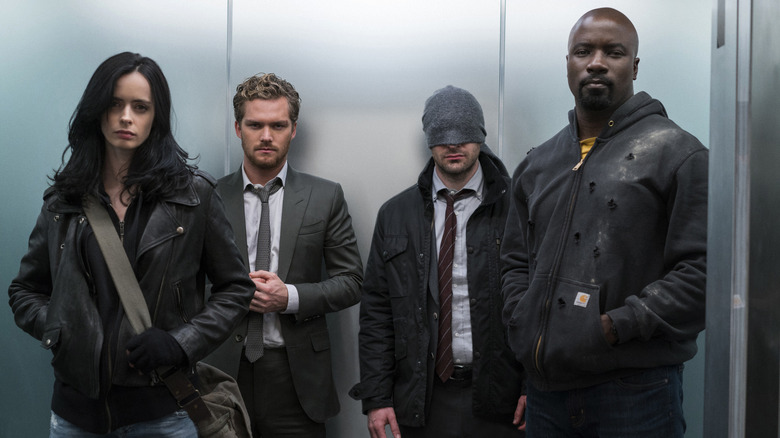 Krysten Ritter as Jessica Jones standing in an elevator with Finn Jones as Danny Rand, Charlie Cox as Matt Murdock, and Mike Colter as Luke Cage in The Defenders