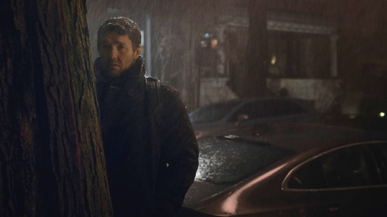 Joel Edgerton as Jason Dessen standing behind a tree trunk on a residential street in the rain in Dark Matter