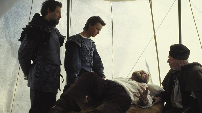 Three men dressed in Middle Ages garb gather around a man writhing on a cot in pain in Flesh+Blood