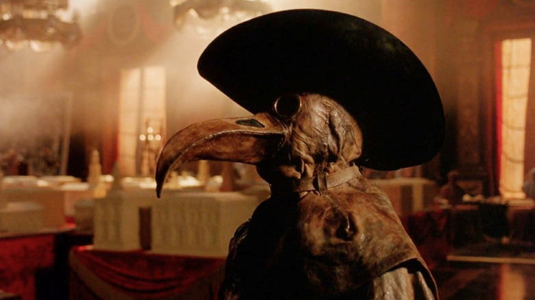 A person dressed in a plague doctor outfit stands in a room bathed in orange light in Restoration