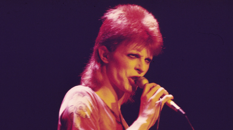 डेव्हिड बोवी लाल प्रकाशात मायक्रोफोन धरून आहे