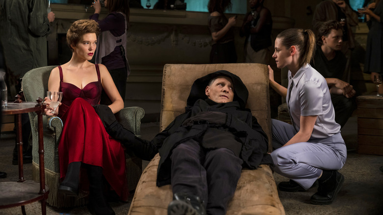 lea seydoux sitting in a red dress next to viggo mortensen laying on a lounge chair with kristen stewart standing on his other side in the movie crimes of the future