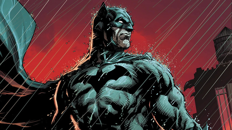 Batman stands in the rain, the night red behind him