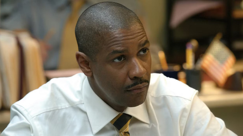 Denzel Washington as Detective Keith Frazier, wearing a white collared shirt and tie, with a focused expression, stands in front of blurred offices in the background of Inside Man (2006).