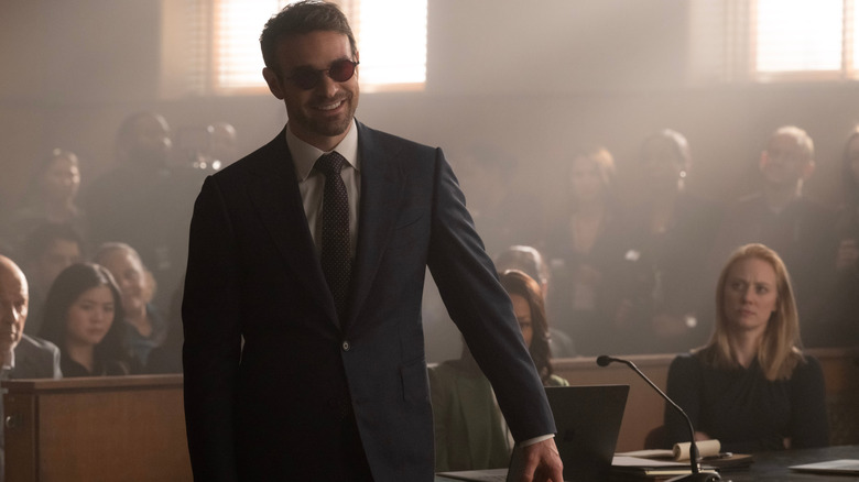 Charlie Cox as Matt Murdock wearing a suit and tie and standing in a courtroom in front of Deborah Ann Woll as Karen Page in Daredevil: Born Again