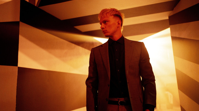 Cyrus wears a suit while standing in a room with geometric wall designs and yellow backlighting in Onslaught