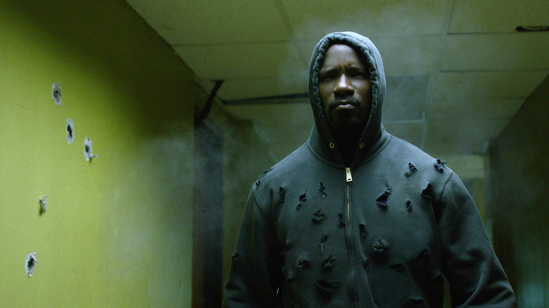 man in grey sweatshirt covered in bullet holes walking down a yellow hallway with bullet holes in the walls