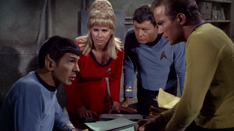 Spock seated at an old 20th century table with Yeoman Rand, McCoy and Kirk standing above him in Star Trek
