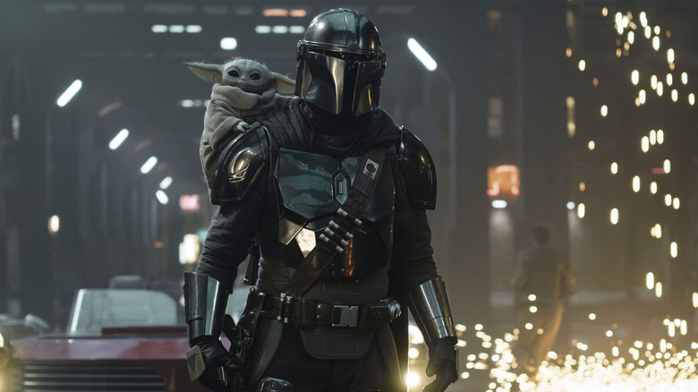 Din Djarin walking past some sparks with Grogu on his back in The Mandalorian and Grogu