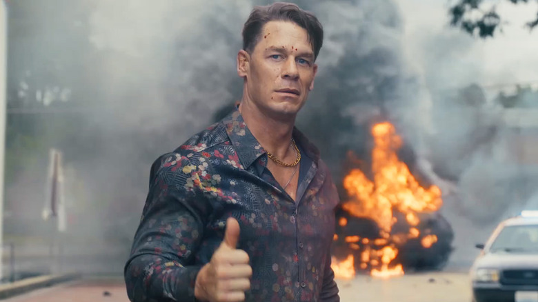 Chris Smith (John Cena) giving a thumbs up with an explosion behind him in a city street in Peacemaker season 2