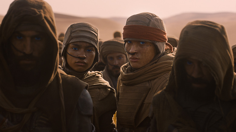 Ida Brooke as Ghanima standing next to Nakota-Wolf Momoa as Leto II in a desert surrounded by several people dressed in robes in Dune: Part Three