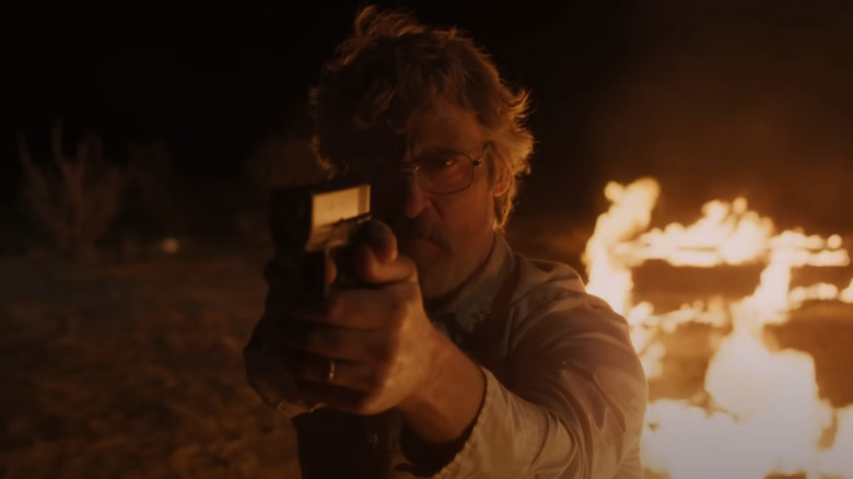 Joe Cross (Joaquin Phoenix) points a gun as fire lights up night in the desert behind him in Eddington