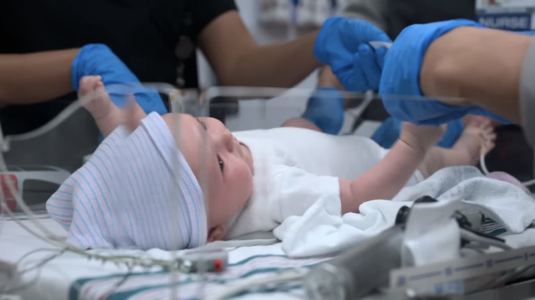 Een baby ligt op zijn rug terwijl de handen van de dokter eraan werken in The Pitt