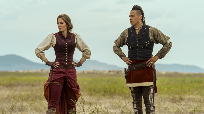 Cornelia and Eli in traveling clothing with their hands on their hips on a plain in The English