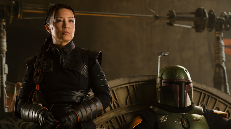 Fennec Shand standing beside a seated Boba Fett, helmet on, in The Book of Boba Fett
