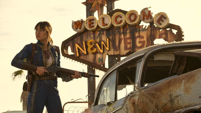 Lucy looks angry and holds a shotgun in front of a ruined Welcome to Vegas sign in Fallout