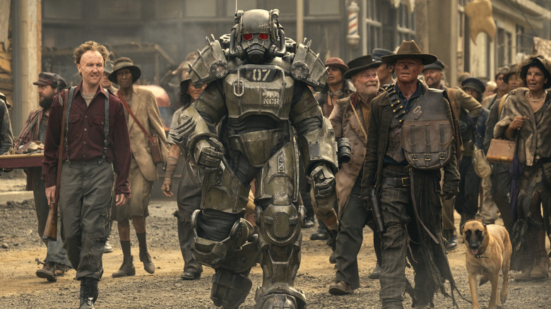 Johnny Pemberton as Thaddeus and Walton Goggins as the Ghoul walking through a crowded street alongside Aaron Moten as Maximus suited up in his armor in Fallout