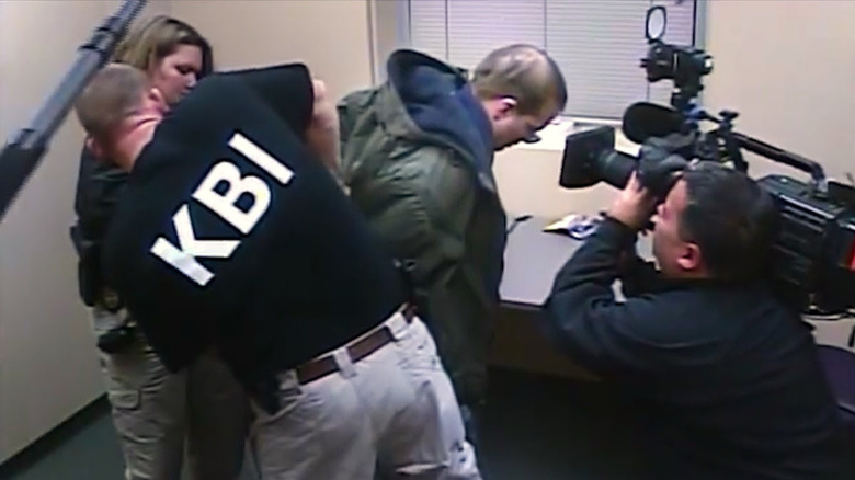 A camera crew films a man being busted for a crime in a beige room in Predators