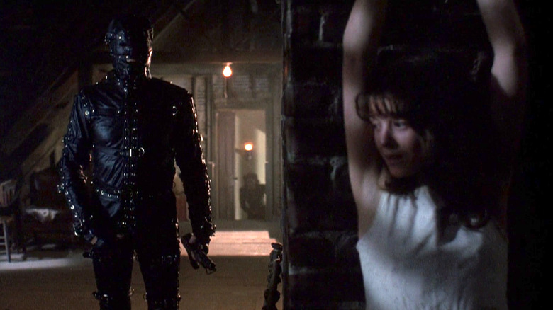 A man in a full black leather gimp suit is looking for a young girl hiding behind a brick wall.