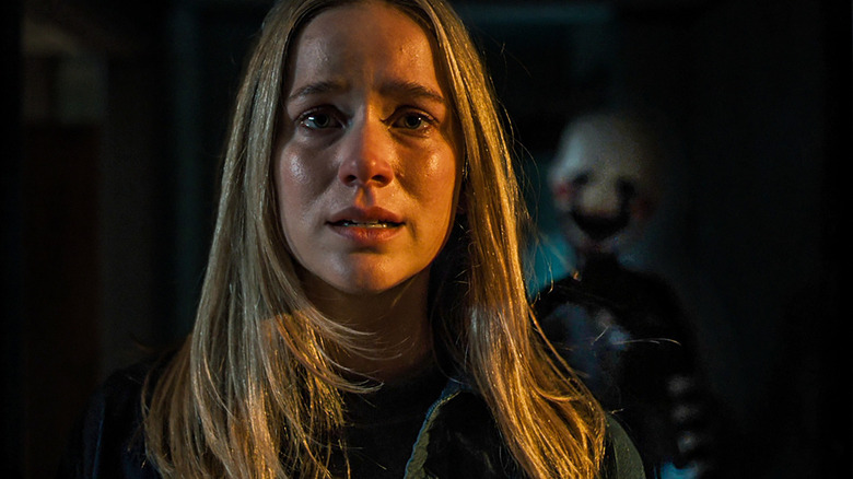 Elizabeth Lail as Vanessa standing in a dark hallway as something creeps up behind her in Five Nights at Freddy's 2