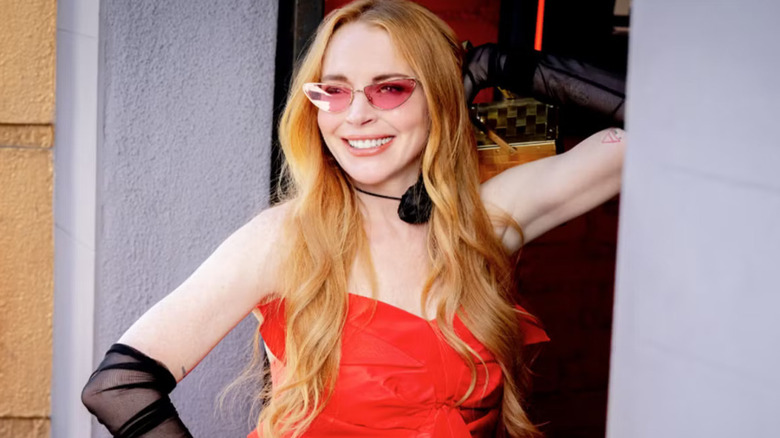 Anna (Lindsay Lohan) wears a strapless red dress with sheer black gloves and pink-tinted cat-eye sunglasses, smiling confidently with one hand on her head and the other on her hip in Freakier Friday