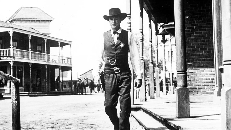 Gary Cooper as Marshal Will Kane, wearing a black cowboy hat, a vest and shirt, and a marshal's star, holds a pistol in his right hand and stands in the main thoroughfare of a small town