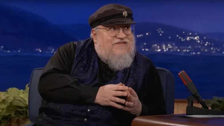 George R.R. Martin sitting on a chair during a talk show appearance with Conan O'Brien