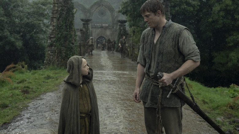 Dexter Sol Ansell as Egg wearing a hood and standing on a road in the rain next to Peter Claffey as Ser Duncan the Tall in A Knight of the Seven Kingdoms
