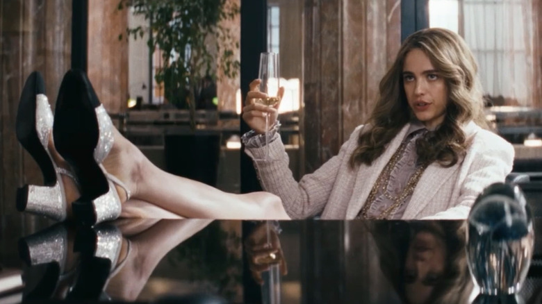 Margaret Qualley's How to Make a Killing character sitting at a desk with her feet up, drinking champagne