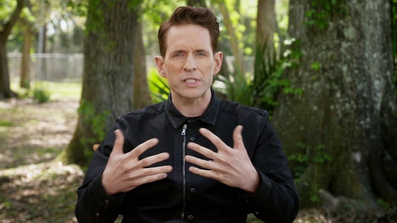 Richard (Glenn Howerton) in a black jacket speaks expressively with his hands raised, in the middle of moss-covered trees in The Hunt