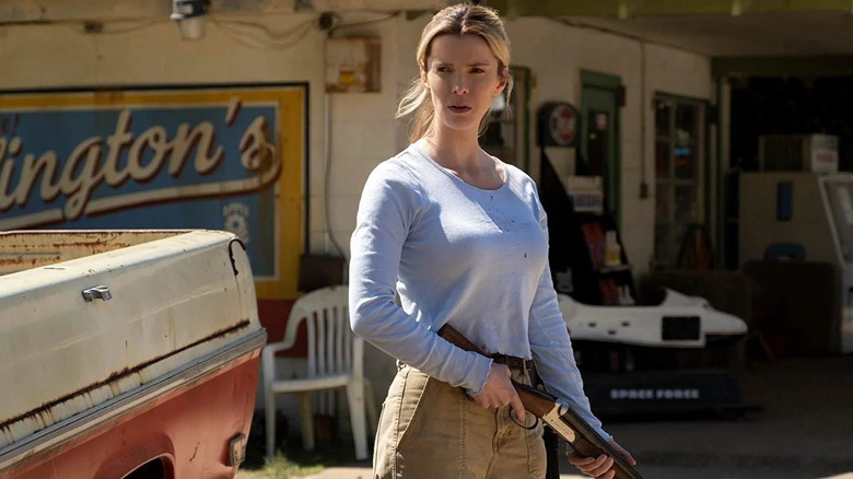 Crystal (Betty Gilpin) stands in front of a rustic roadside store next to a dirty truck, with a serious expression while holding a shotgun in The Hunt