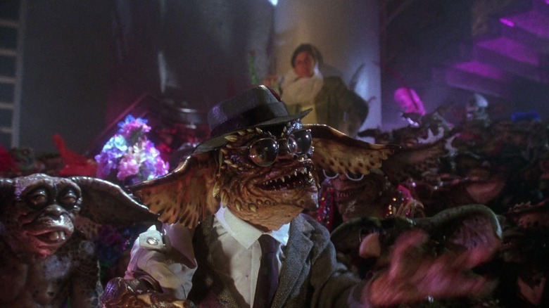 Brainy Gremlin, wearing a hat and glasses while dressed in a formal suit and tie, addresses the partying crowd of gremlins in "Gremlins 2: The New Batch"