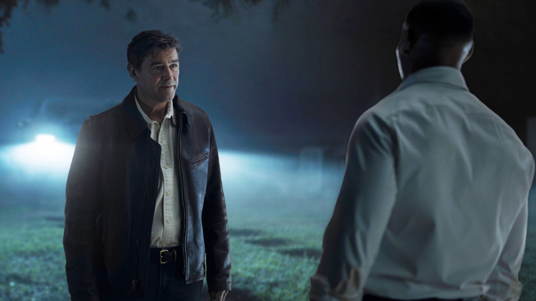 Kyle Chandler's Hal Jordan sits opposite Aaron Pierre's John Stewart stand opposite one another in a graveyard in front of a parked car in Lanterns