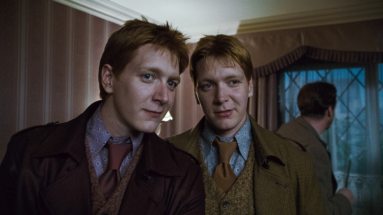 James Phelps as Fred Weasley and Oliver Phelps as George Weasley, with David Thewlis as Remus Lupin in the background, in Harry Potter and the Deathly Hallows: Part 2, standing in a house and looking at something together with slight smirks.