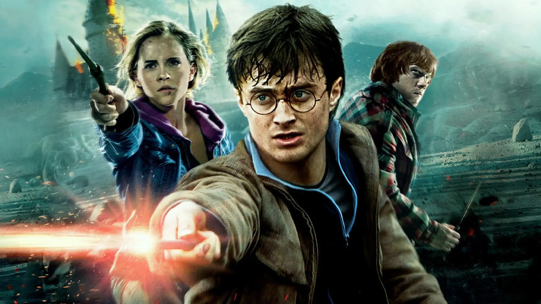 Harry (Daniel Radcliffe), Hermione (Emma Watson), and Ron (Rupert Grint) with wands drawn in front of a battle-damaged Hogwarts in a poster for Harry Potter and the Deathly Hallows Part 2