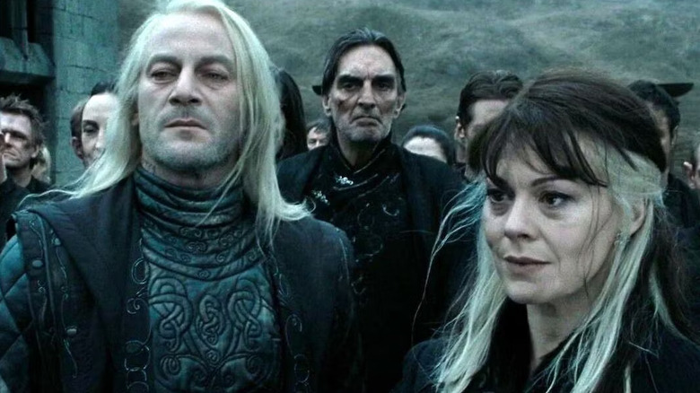 Jason Isaacs as Lucius Malfoy and Helen McCrory as Narcissa Malfoy in Harry Potter in the Deathly Hallows: Part 2, standing next to each other against a large group of Death Eathers, looking with contempt off at someone out of frame.