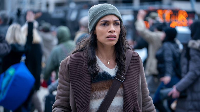 Rosario Dawson as Zee wearing a beanie and surrounded by people marching in the background in DMZ