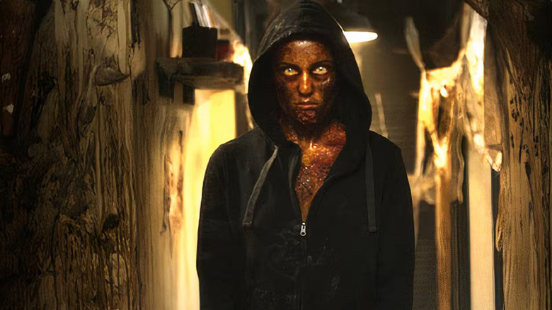 An infected person stands in a dingy basement, staring intensely in a black hooded jacket, their yellowed skin covered in red bumps and wide eyes glowing yellow in Hidden