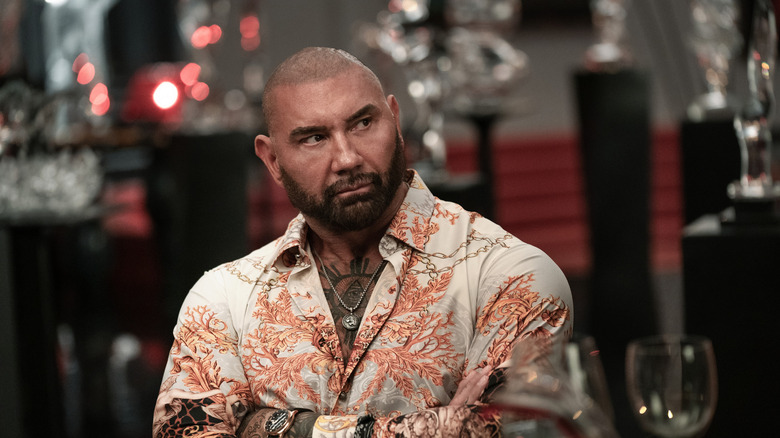 Dave Bautista as Duke sitting at the table with his arms crossed in Glass Onion: A Knives Out Mystery